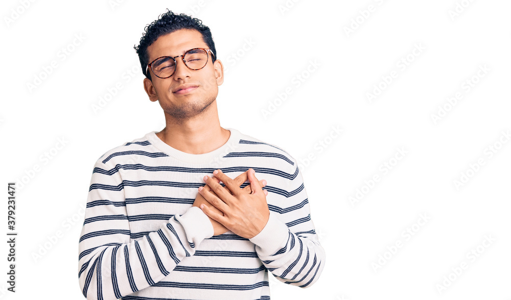 Hispanic handsome young man wearing casual clothes and glasses smiling with hands on chest with closed eyes and grateful gesture on face. health concept. Hispanic handsome young man wearing casual clothes and glasses smiling with hands on chest with closed eyes and grateful gesture on face. health concept.