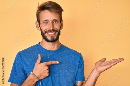 Handsome caucasian man with beard wearing casual clothes amazed and smiling to the camera while presenting with hand and pointing with finger.
