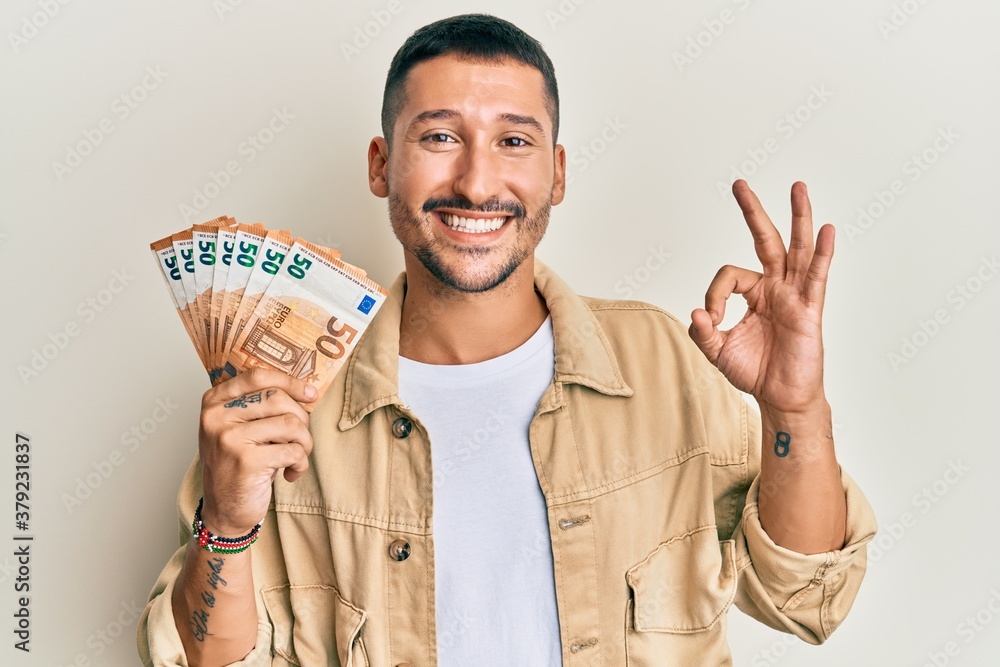 Handsome man with tattoos holding 50 euro banknotes doing ok sign with fingers, smiling friendly gesturing excellent symbol - obrazy, fototapety, plakaty 