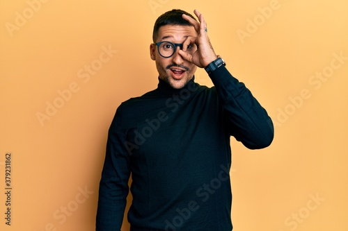 Handsome man with tattoos wearing turtleneck sweater and glasses doing ok gesture shocked with surprised face, eye looking through fingers. unbelieving expression. © Krakenimages.com