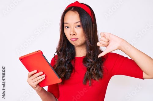 Young beautiful chinese girl holding touchpad with angry face, negative sign showing dislike with thumbs down, rejection concept