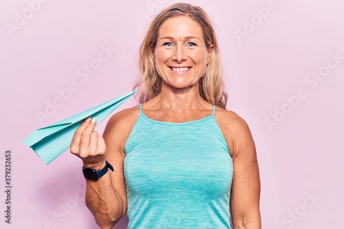 Middle age caucasian blonde woman holding paper airplane looking positive and happy standing and smiling with a confident smile showing teeth