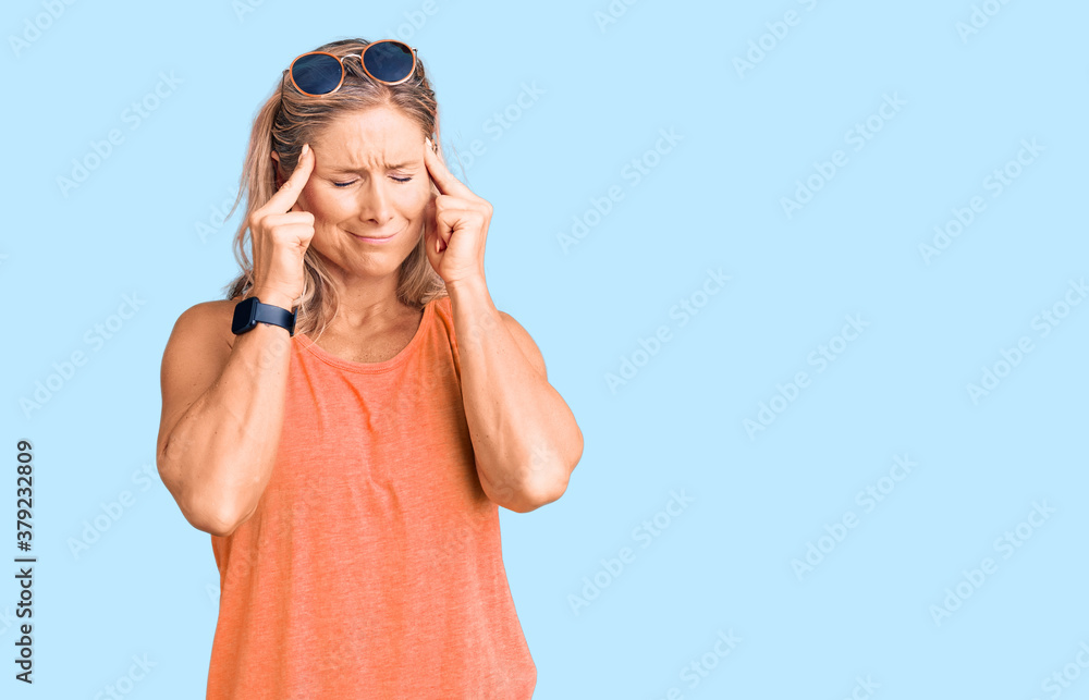 Middle age fit blonde woman wearing casual summer clothes and sunglasses with hand on head, headache because stress. suffering migraine. Middle age fit blonde woman wearing casual summer clothes and sunglasses with hand on head, headache because stress. suffering migraine.