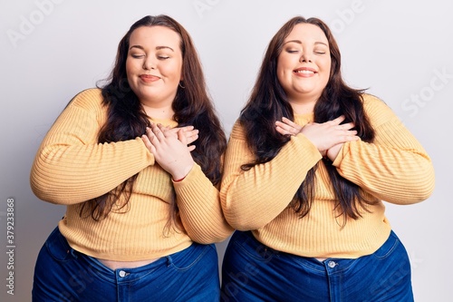Young plus size twins wearing casual clothes smiling with hands on chest with closed eyes and grateful gesture on face. health concept.
