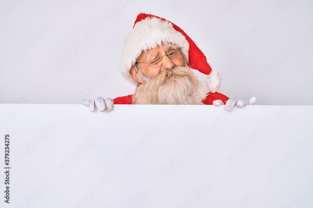 Old senior man with grey hair and long beard wearing santa claus costume holding banner winking looking at the camera with sexy expression, cheerful and happy face. Old senior man with grey hair and long beard wearing santa claus costume holding banner winking looking at the camera with sexy expression, cheerful and happy face.