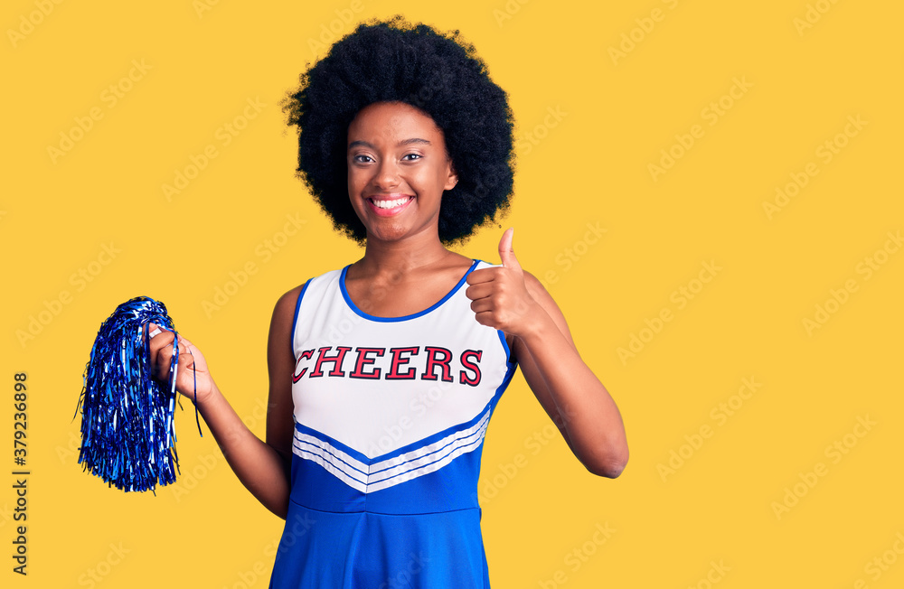 Young african american woman wearing cheerleader uniform holding pompom doing happy thumbs up gesture with hand. approving expression looking at the camera showing success. Young african american woman wearing cheerleader uniform holding pompom doing happy thumbs up gesture with hand. approving expression looking at the camera showing success.