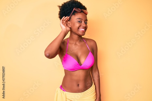 Young african american woman wearing bikini smiling with hand over ear listening an hearing to rumor or gossip. deafness concept.