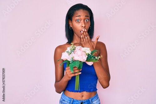 Young african american woman holding flowers covering mouth with hand, shocked and afraid for mistake. surprised expression