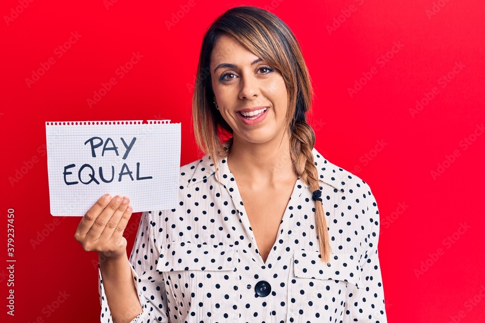 Young beautiful woman holding pay equal message paper looking positive and happy standing and smiling with a confident smile showing teeth Young beautiful woman holding pay equal message paper looking positive and happy standing and smiling with a confident smile showing teeth