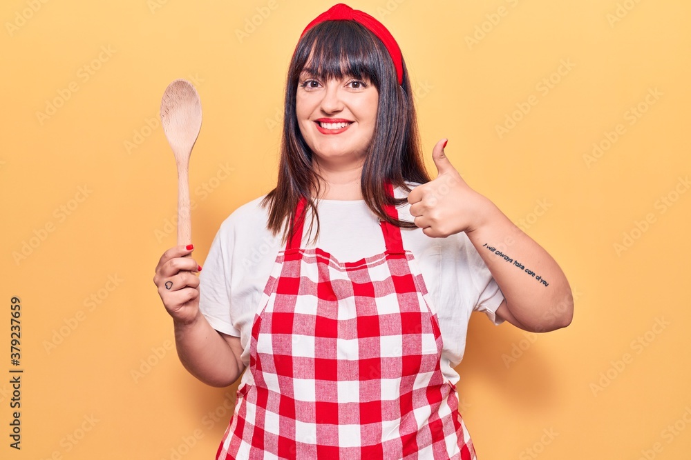 Young plus size woman wearing apron holding wooden spoon smiling happy and positive, thumb up doing excellent and approval sign Young plus size woman wearing apron holding wooden spoon smiling happy and positive, thumb up doing excellent and approval sign