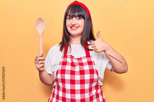 Young plus size woman wearing apron holding wooden spoon smiling happy and positive, thumb up doing excellent and approval sign