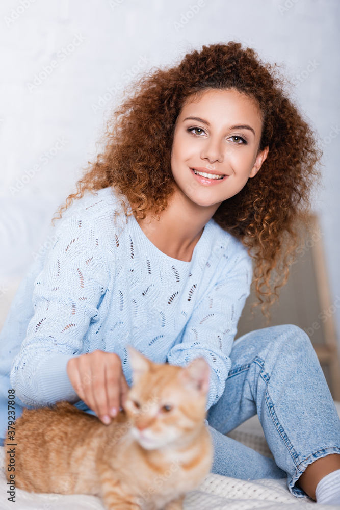 Selective focus of red haired woman in sweater looking at camera near cat at home