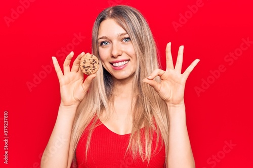 Young beautiful blonde woman holding cookie doing ok sign with fingers, smiling friendly gesturing excellent symbol