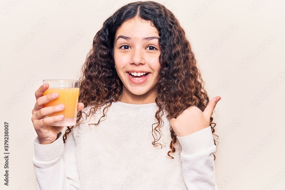 Beautiful kid girl with curly hair drinking glass of orange juice pointing thumb up to the side smiling happy with open mouth Beautiful kid girl with curly hair drinking glass of orange juice pointing thumb up to the side smiling happy with open mouth