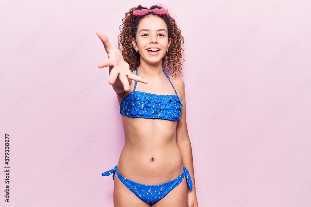 Naklejka premium Beautiful kid girl with curly hair wearing bikini and sunglasses smiling friendly offering handshake as greeting and welcoming. successful business.