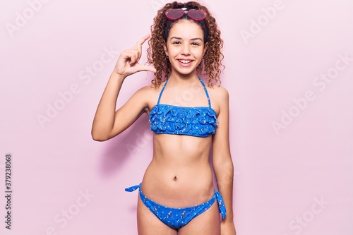 Beautiful kid girl with curly hair wearing bikini and sunglasses smiling and confident gesturing with hand doing small size sign with fingers looking and the camera. measure concept.