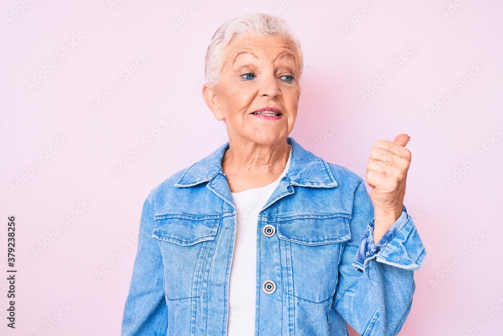 Senior beautiful woman with blue eyes and grey hair wearing casual denim jacket smiling with happy face looking and pointing to the side with thumb up. Senior beautiful woman with blue eyes and grey hair wearing casual denim jacket smiling with happy face looking and pointing to the side with thumb up.