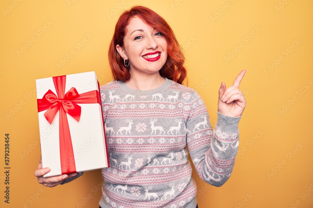 Young beautiful redhead woman holding christmas gift over isolated yellow background smiling happy pointing with hand and finger to the side Young beautiful redhead woman holding christmas gift over isolated yellow background smiling happy pointing with hand and finger to the side