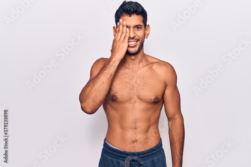 Young latin man standing shirtless covering one eye with hand  confident smile on face and surprise emotion.