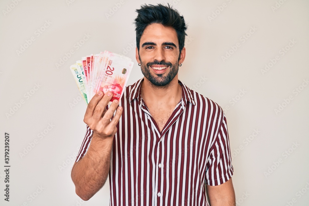 Handsome hispanic man with beard holding israel shekels looking positive and happy standing and smiling with a confident smile showing teeth Handsome hispanic man with beard holding israel shekels looking positive and happy standing and smiling with a confident smile showing teeth