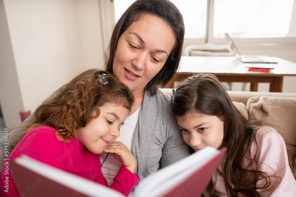 Lovely latin mother sitting in sofa reading for her kids. Indoors at home living room. Togetherness, multi-generation family, support concept. Lovely latin mother sitting in sofa reading for her kids. Indoors at home living room. Togetherness, multi-generation family, support concept.
