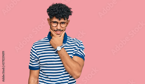Young arab man wearing casual clothes and glasses looking confident at the camera smiling with crossed arms and hand raised on chin. thinking positive. © Krakenimages.com