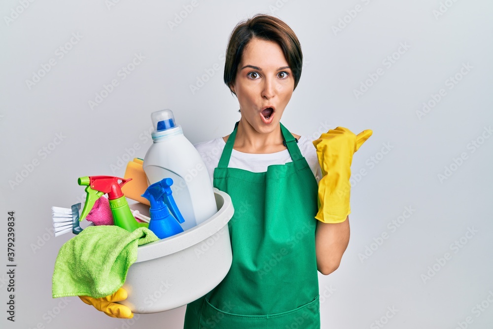 Young brunette woman with short hair wearing apron holding cleaning products surprised pointing with hand finger to the side, open mouth amazed expression. Young brunette woman with short hair wearing apron holding cleaning products surprised pointing with hand finger to the side, open mouth amazed expression.