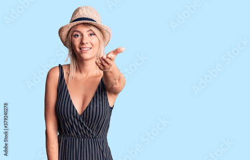 Young beautiful blonde woman wearing summer hat and dress smiling friendly offering handshake as greeting and welcoming. successful business.