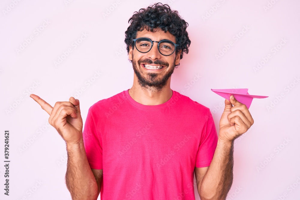 Handsome young man with curly hair and bear holding paper airplane smiling happy pointing with hand and finger to the side Handsome young man with curly hair and bear holding paper airplane smiling happy pointing with hand and finger to the side