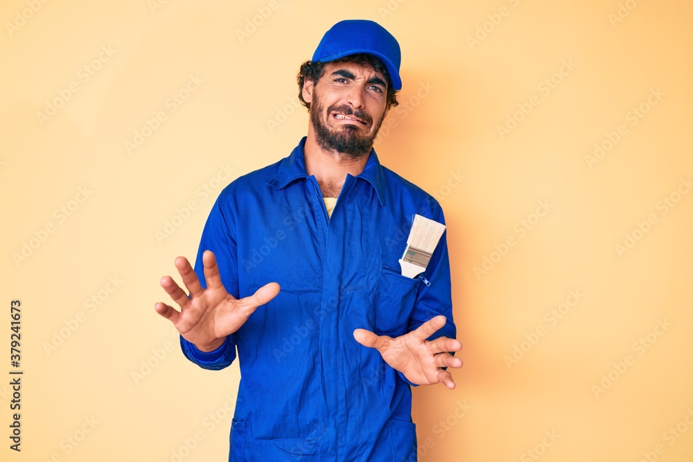 Handsome young man with curly hair and bear wearing builder jumpsuit uniform disgusted expression, displeased and fearful doing disgust face because aversion reaction. with hands raised