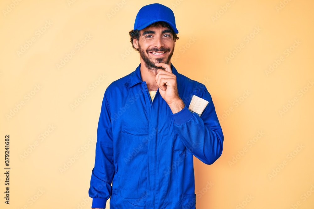 Handsome young man with curly hair and bear wearing builder jumpsuit uniform looking confident at the camera with smile with crossed arms and hand raised on chin. thinking positive.
