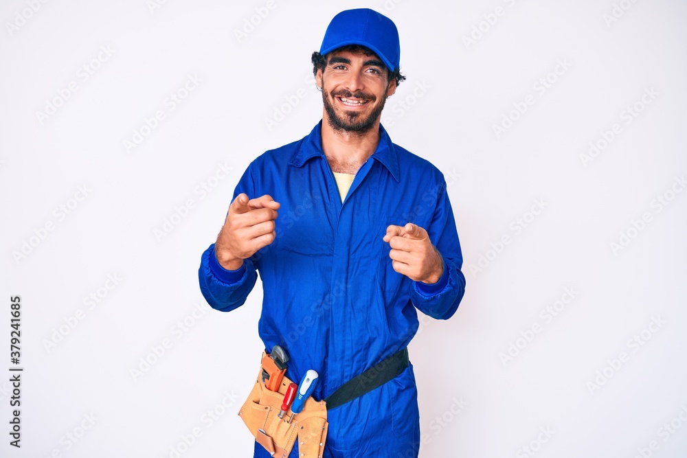 Handsome young man with curly hair and bear weaing handyman uniform pointing fingers to camera with happy and funny face. good energy and vibes.