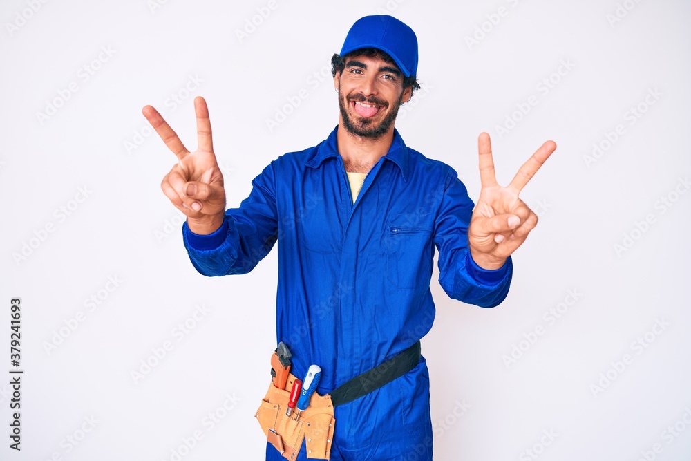 Handsome young man with curly hair and bear weaing handyman uniform smiling with tongue out showing fingers of both hands doing victory sign. number two.