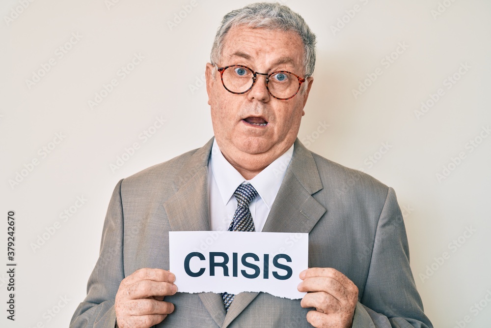 Senior grey-haired man wearing business suit holding crisis paper clueless and confused expression. doubt concept. Senior grey-haired man wearing business suit holding crisis paper clueless and confused expression. doubt concept.