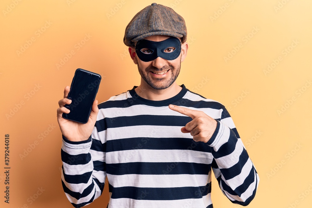 Young handsome bald man wearing burglar mask holding smartphone smiling happy pointing with hand and finger Young handsome bald man wearing burglar mask holding smartphone smiling happy pointing with hand and finger