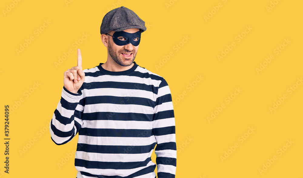 Young handsome man wearing burglar mask pointing finger up with successful idea. exited and happy. number one. Young handsome man wearing burglar mask pointing finger up with successful idea. exited and happy. number one.