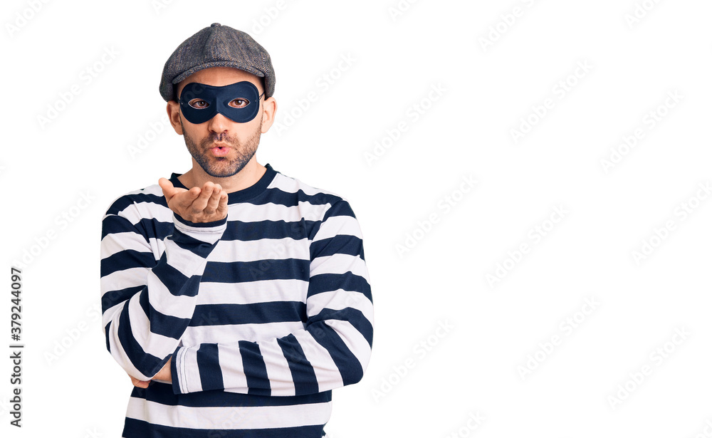 Young handsome man wearing burglar mask looking at the camera blowing a kiss with hand on air being lovely and sexy. love expression. Young handsome man wearing burglar mask looking at the camera blowing a kiss with hand on air being lovely and sexy. love expression.