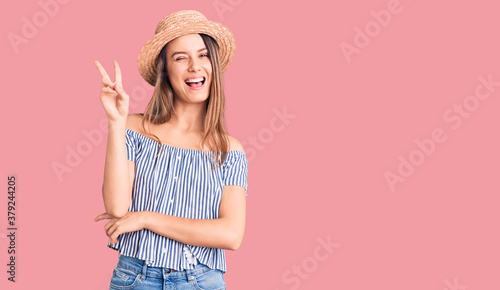Young beautiful girl wearing hat and t shirt smiling with happy face winking at the camera doing victory sign. number two.