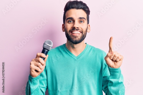 Young handsome man with beard singing song using microphone smiling with an idea or question pointing finger with happy face, number one