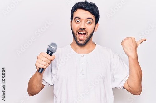 Young hispanic man singing song using microphone pointing thumb up to the side smiling happy with open mouth