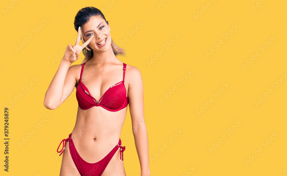 Fototapeta premium Young beautiful woman wearing bikini doing peace symbol with fingers over face, smiling cheerful showing victory