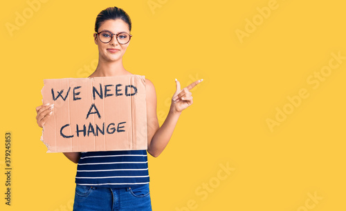Young beautiful woman holding we need a change banner smiling happy pointing with hand and finger to the side
