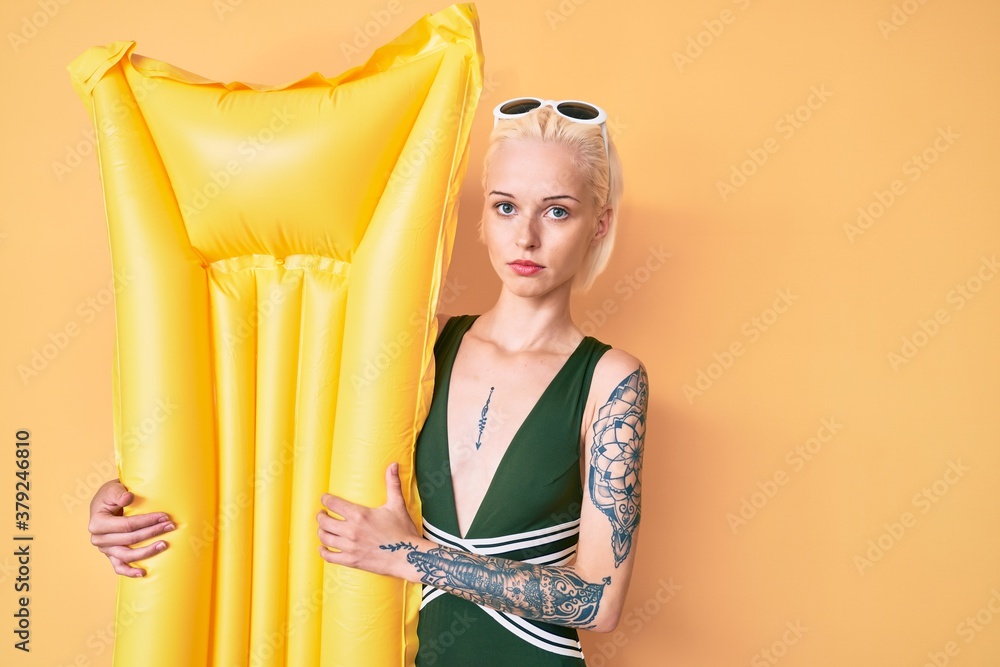 Obraz premium Young blonde woman with tattoo wearing swimsuit and holding summer matress float thinking attitude and sober expression looking self confident