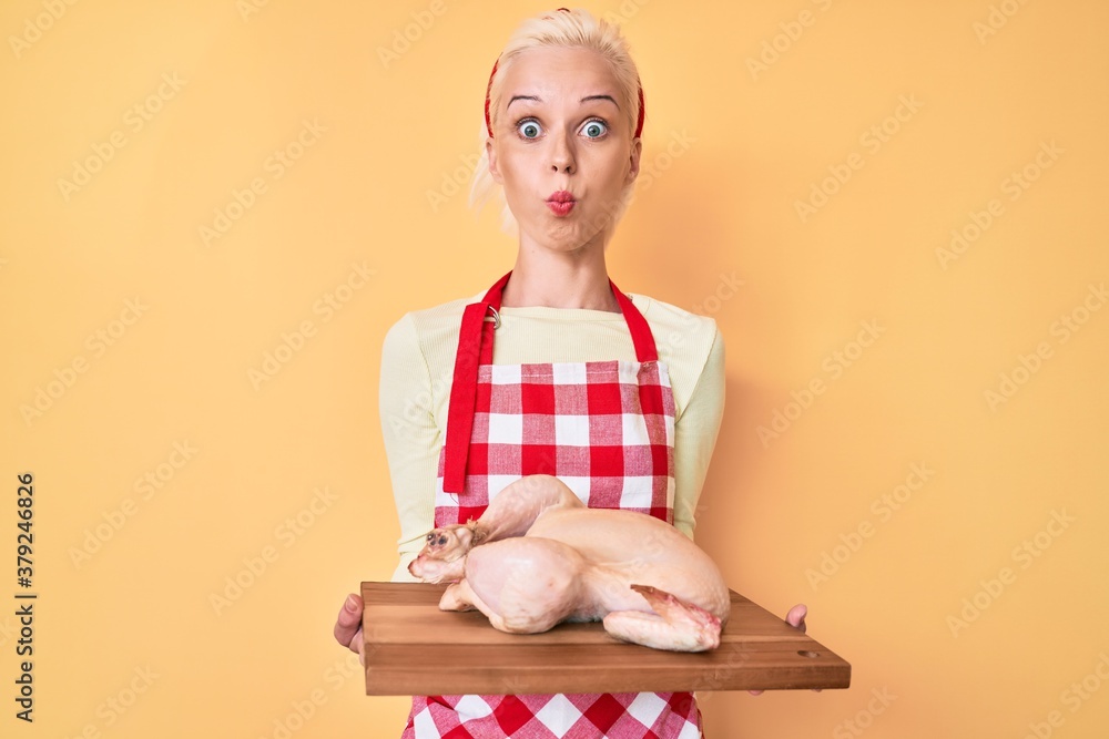 Young blonde woman wearing apron cooking chicken puffing cheeks with funny face. mouth inflated with air, catching air. Young blonde woman wearing apron cooking chicken puffing cheeks with funny face. mouth inflated with air, catching air.