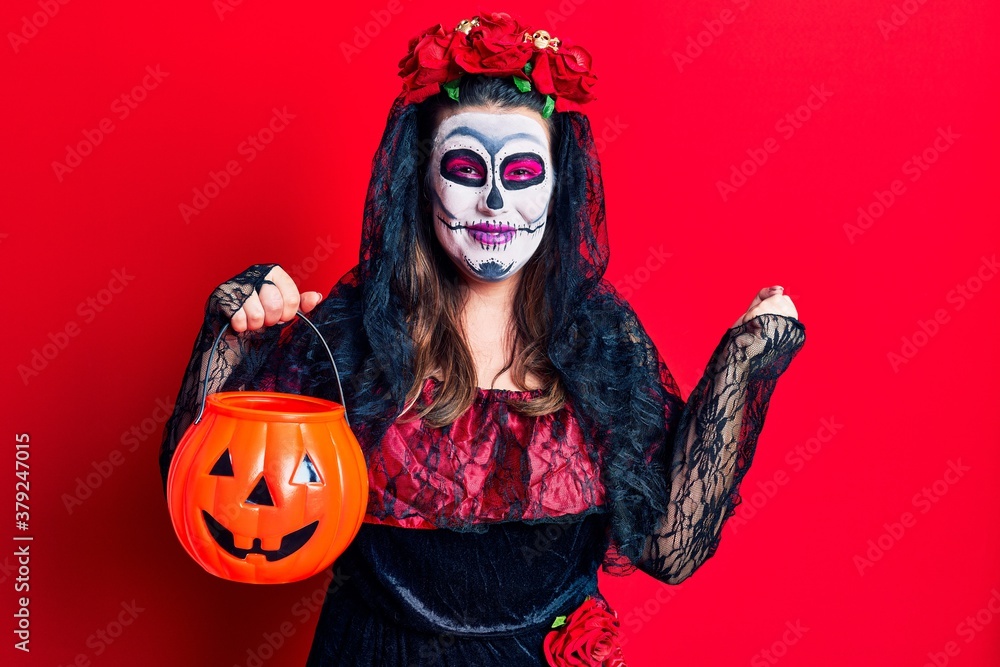 Young woman wearing day of the dead costume holding pumpkin screaming proud, celebrating victory and success very excited with raised arms