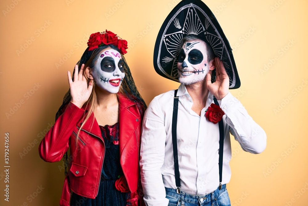 Couple wearing day of the dead costume over yellow smiling with hand over ear listening an hearing to rumor or gossip. deafness concept. Couple wearing day of the dead costume over yellow smiling with hand over ear listening an hearing to rumor or gossip. deafness concept.