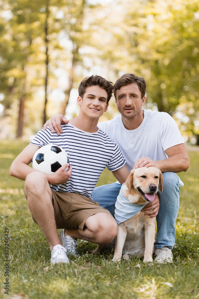 Obraz premium father sitting on grass near teenager son with football and golden retriever