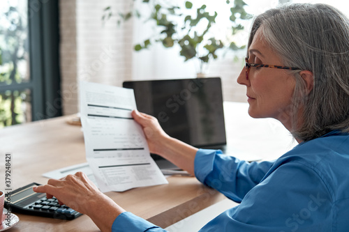 Senior mature business woman holding paper bill using calculator, old lady managing account finances, calculating money budget tax, planning banking loan debt pension payment sit at home office table. photo