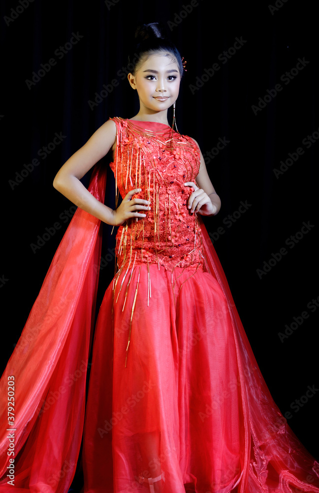 Portrait of a cute Asian schoolgirl wearing a red evening dress and makeup on a black background.