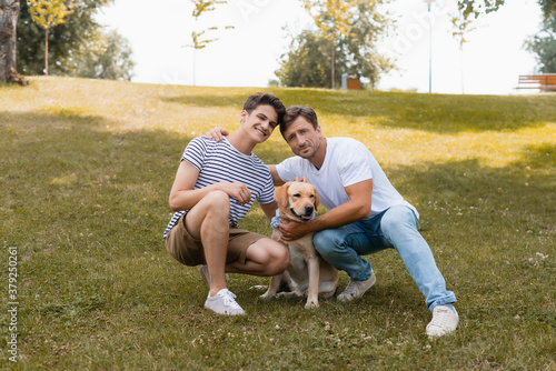 father hugging teenager boy near golden retriever while sitting on grass in park